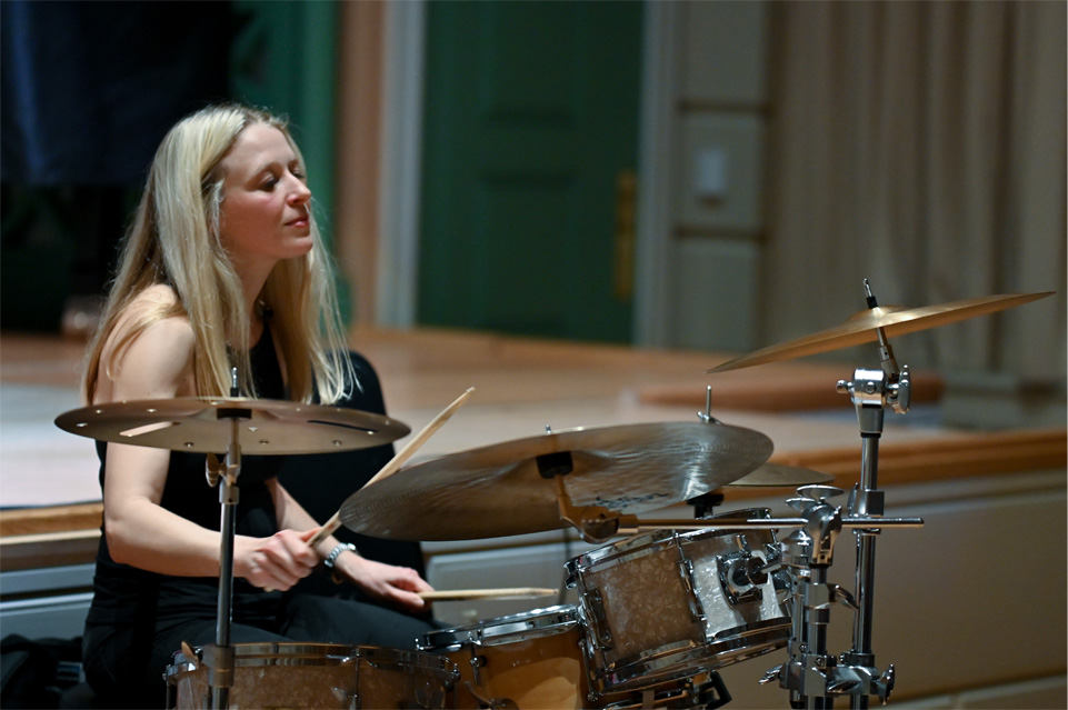 Female drummer, with blonde hair and wearing a black outfit, playing on the drum kit at Amaryllis Fleming Concert Hall.